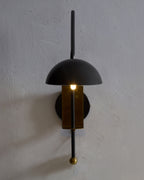 Marcello wall sconce mounted on a textured wall, featuring a black dome shade, vertical black stem, and brass center with warm illuminated bulb.