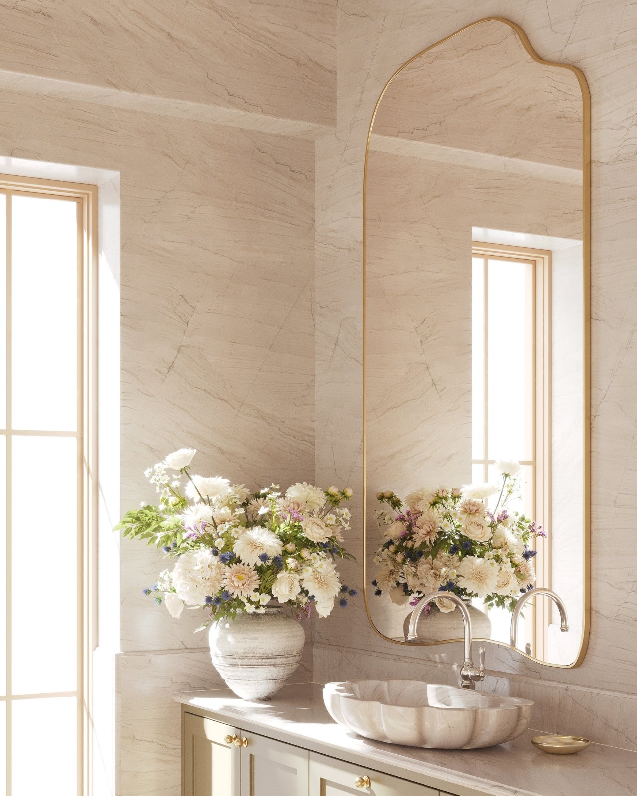 Asymmetrical wall-mounted mirror with a thin brass-colored frame above a vanity, reflecting a marble sink, floral arrangement, and natural daylight from a nearby window in a bright bathroom.