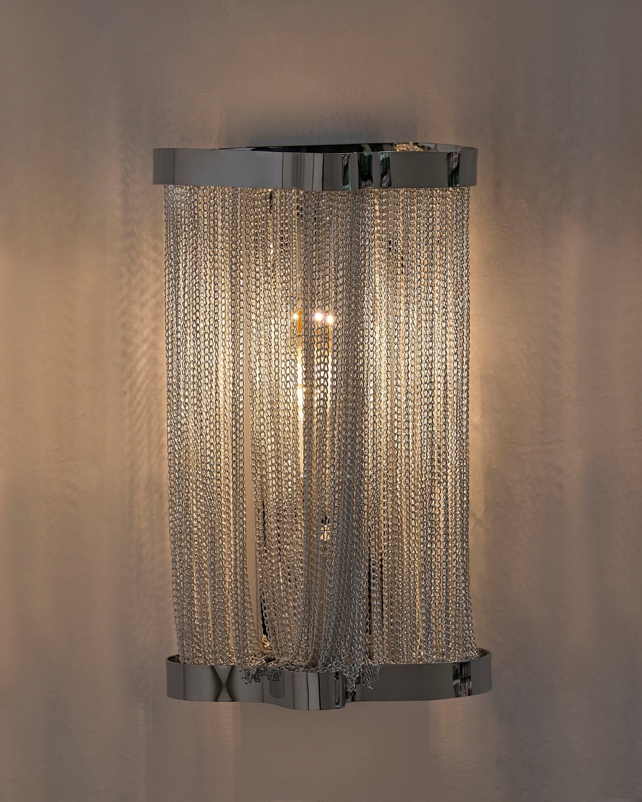 Skulpta Wall Sconce mounted on a wall, featuring a polished silver finish and cascading metal chain curtain that softly diffuses warm light, creating an elegant modern ambiance.