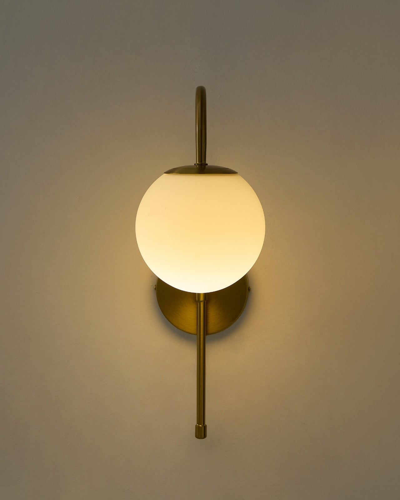 Wall-mounted light fixture with a brass vertical rod, a round brass backplate, and a spherical opaque white glass shade emitting warm light.