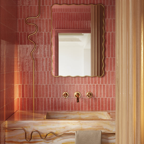 Rectangular wall mirror with a softly wavy gold frame mounted on a pink tiled wall above a marble bathroom sink with brass fixtures.