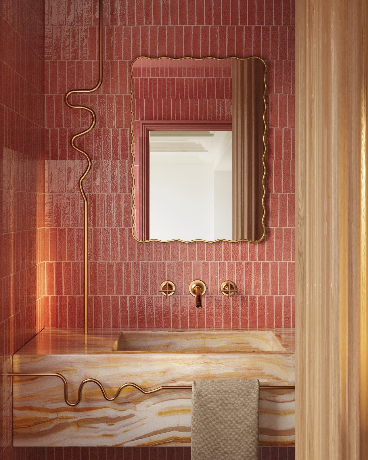 Rectangular wall mirror with a softly wavy gold frame mounted on a pink tiled wall above a marble bathroom sink with brass fixtures.