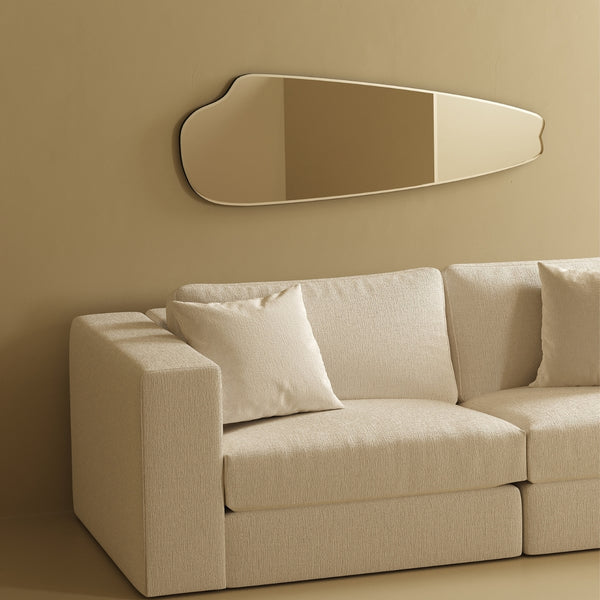 Horizontally mounted frameless wall mirror with an elongated organic shape, displayed above a light-colored sofa in a minimalist interior.