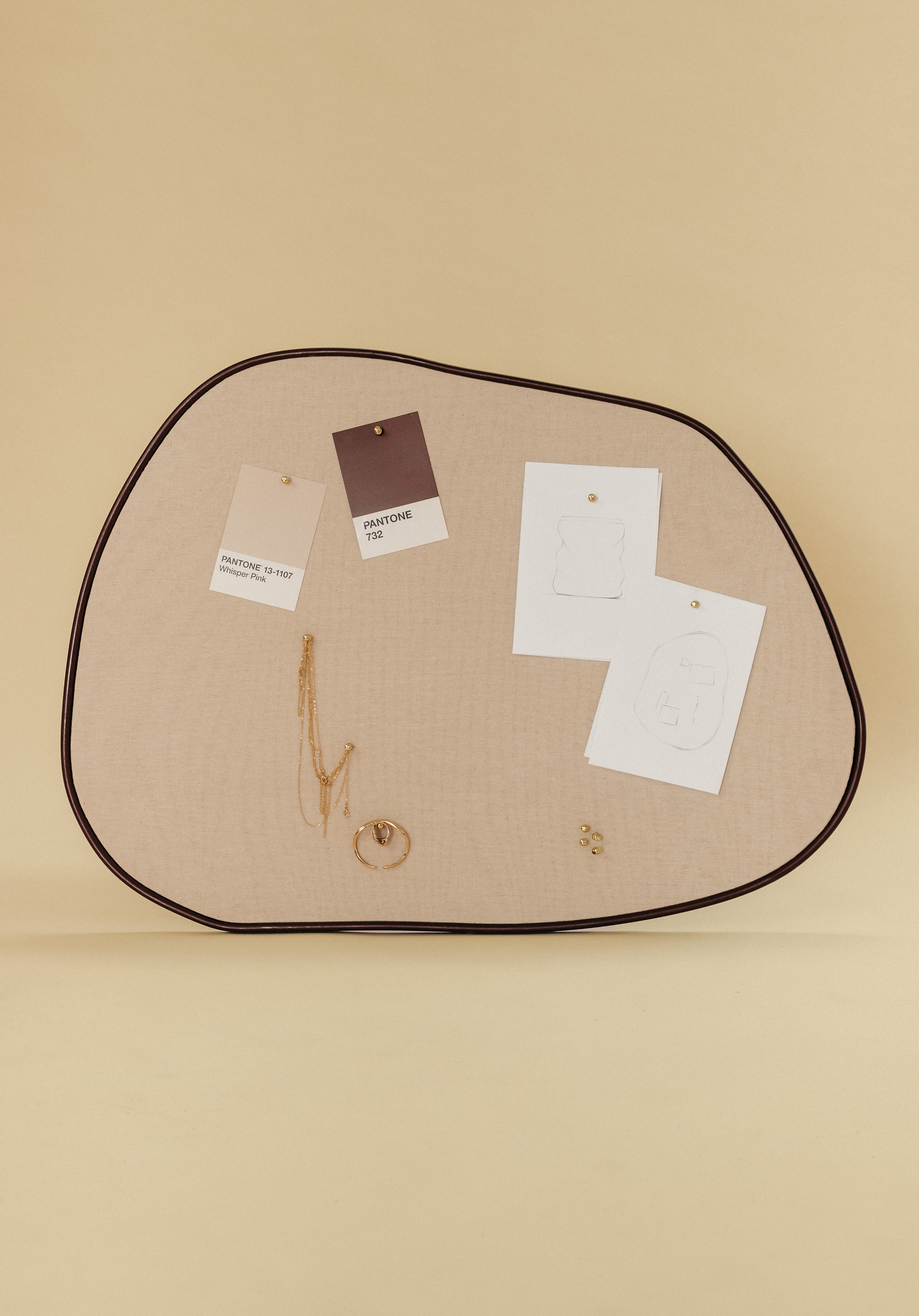 Asymmetrical beige fabric pinboard with a brown outer frame, displaying pinned color cards, sketches, and gold jewelry on a neutral background.