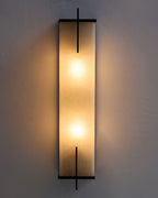 Agatha oversized wall sconce illuminated on a textured wall, featuring a tall alabaster panel that emits a warm, diffused glow, framed by slim black copper accents with soft light cast outward.