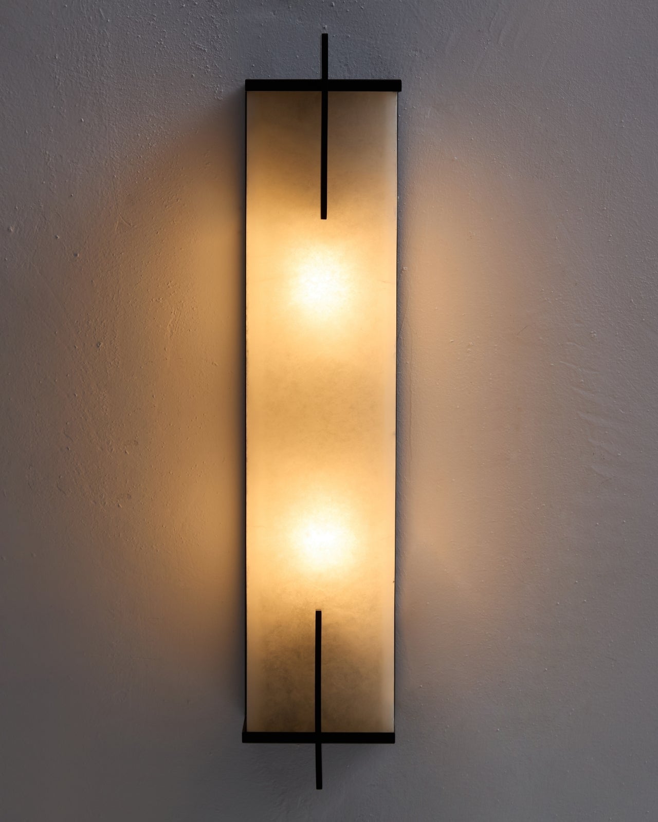 Agatha oversized wall sconce illuminated on a textured wall, featuring a tall alabaster panel that emits a warm, diffused glow, framed by slim black copper accents with soft light cast outward.
