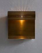 Wall-mounted antique brass light fixture with a perforated horizontal cylindrical shade above a flat rectangular backplate, illuminated against a textured wall.