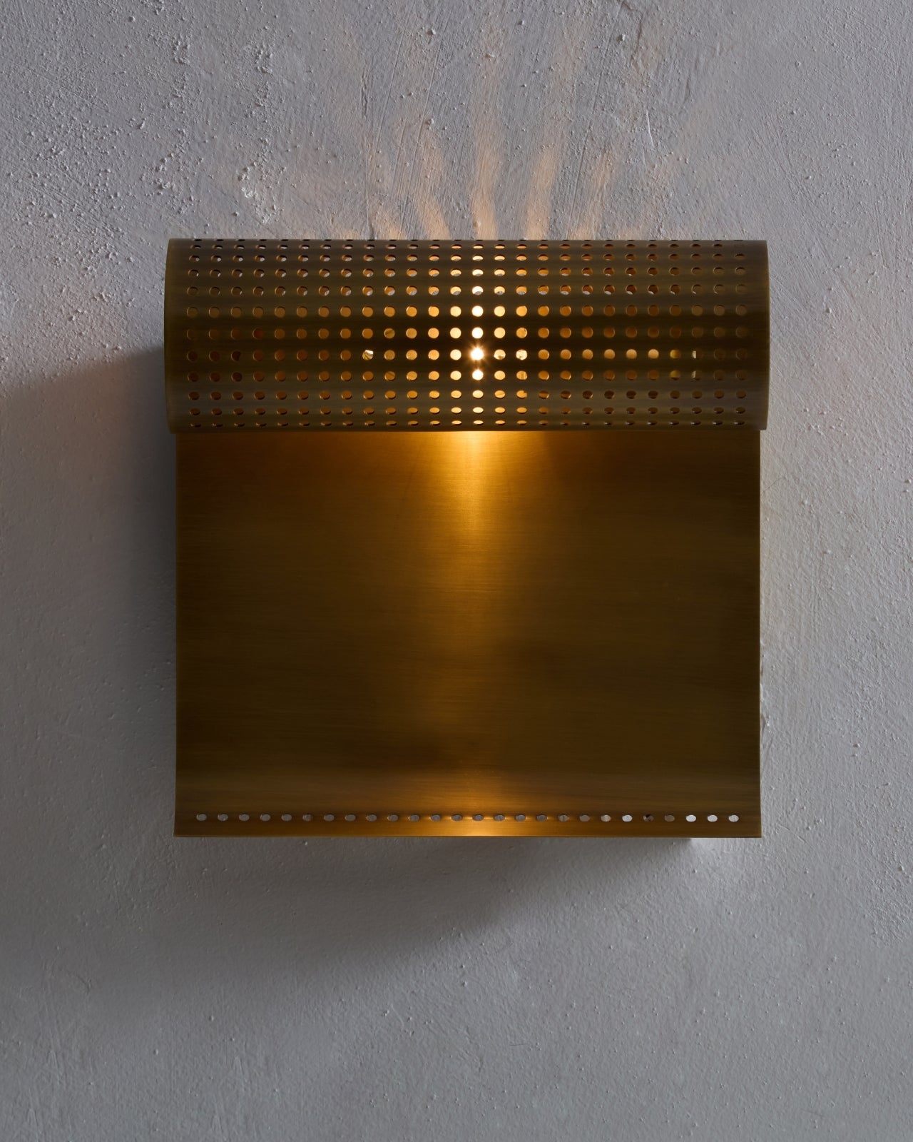 Wall-mounted antique brass light fixture with a perforated horizontal cylindrical shade above a flat rectangular backplate, illuminated against a textured wall.