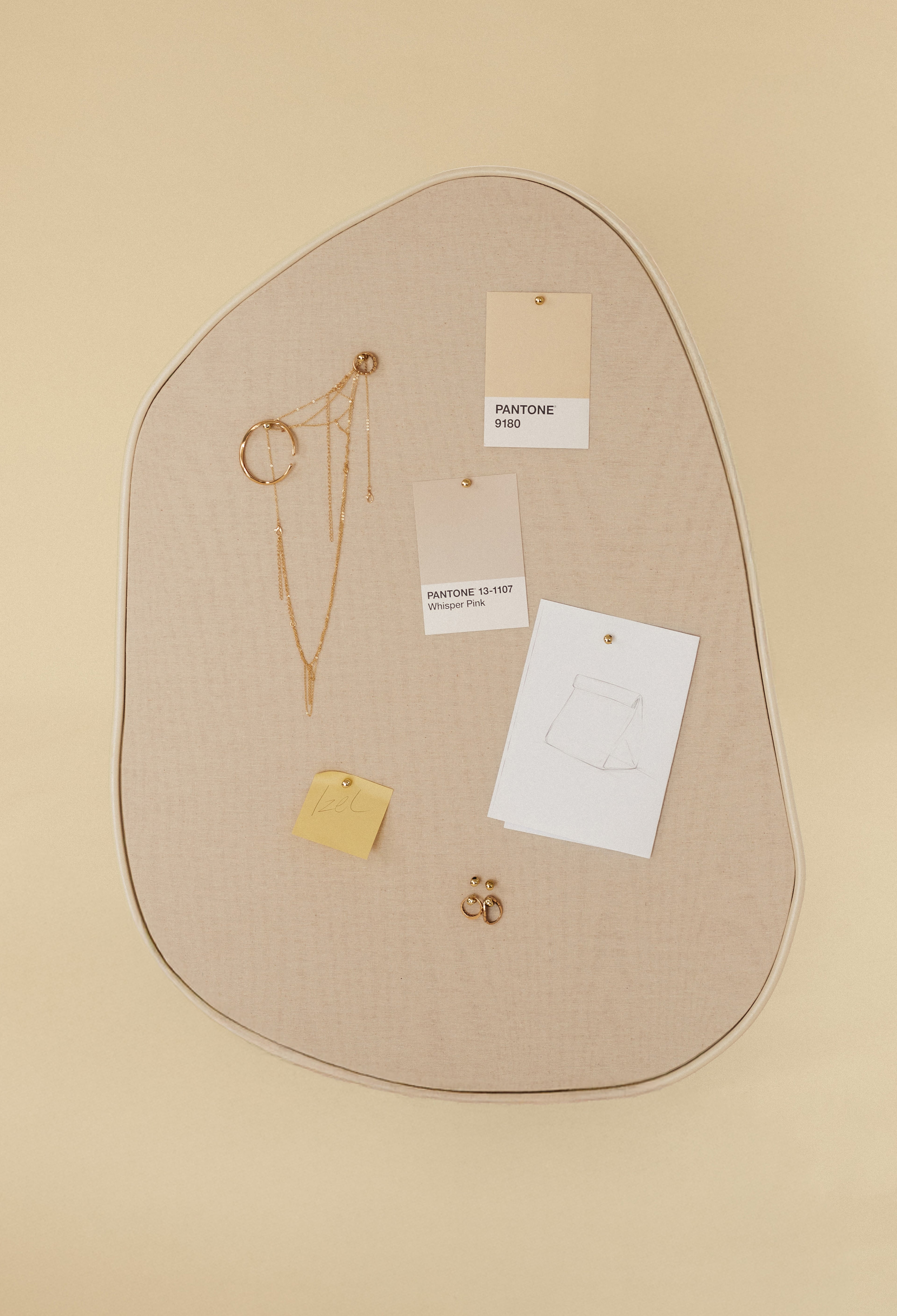 Asymmetrical beige fabric pinboard with a beige outer edge, displaying pinned Pantone color cards, paper sketches, a sticky note, and gold jewelry on a light background.