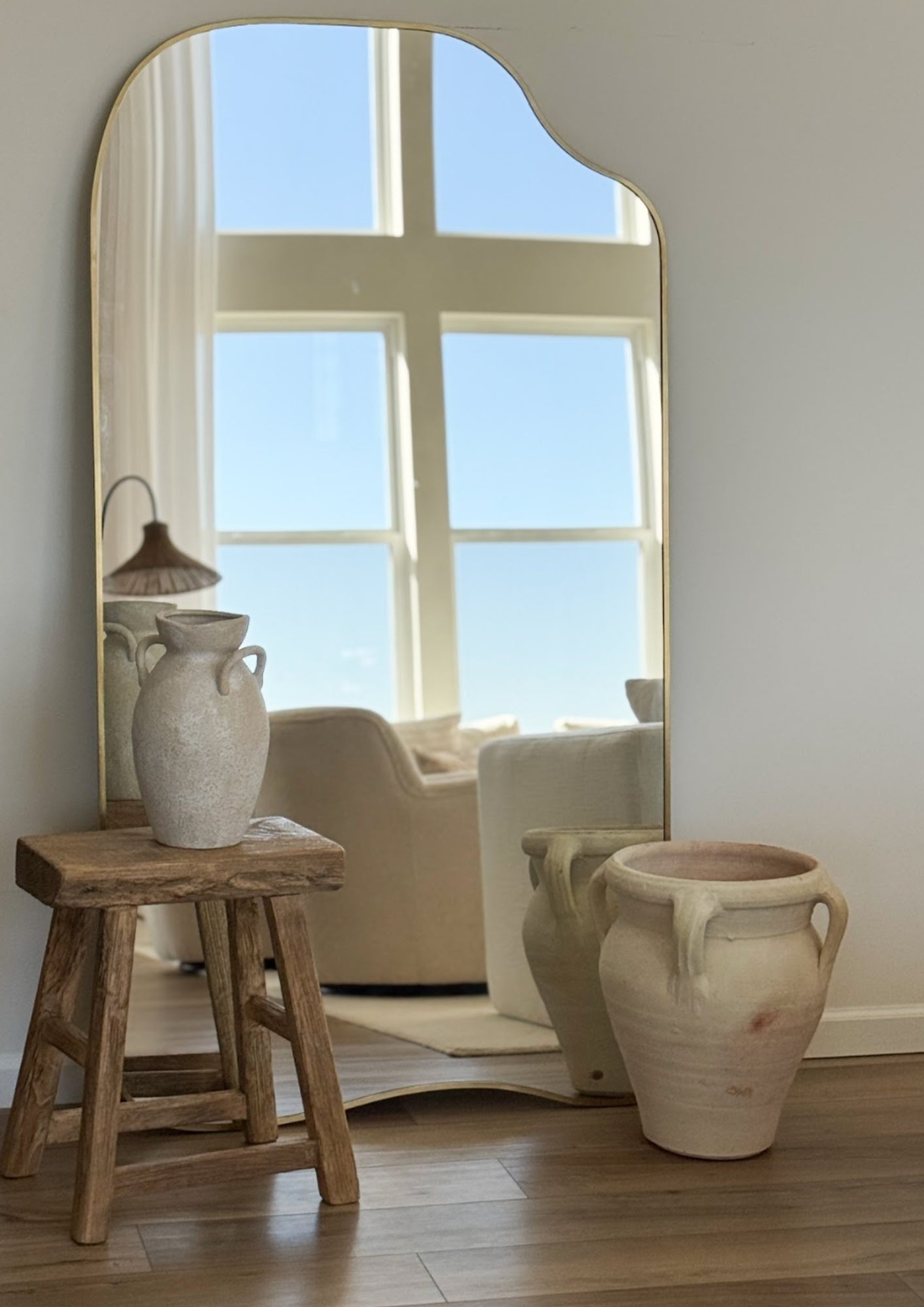 Organic-shaped full length mirror with brass frame, reflecting a sunlit interior with a window, stool, and ceramic vases.