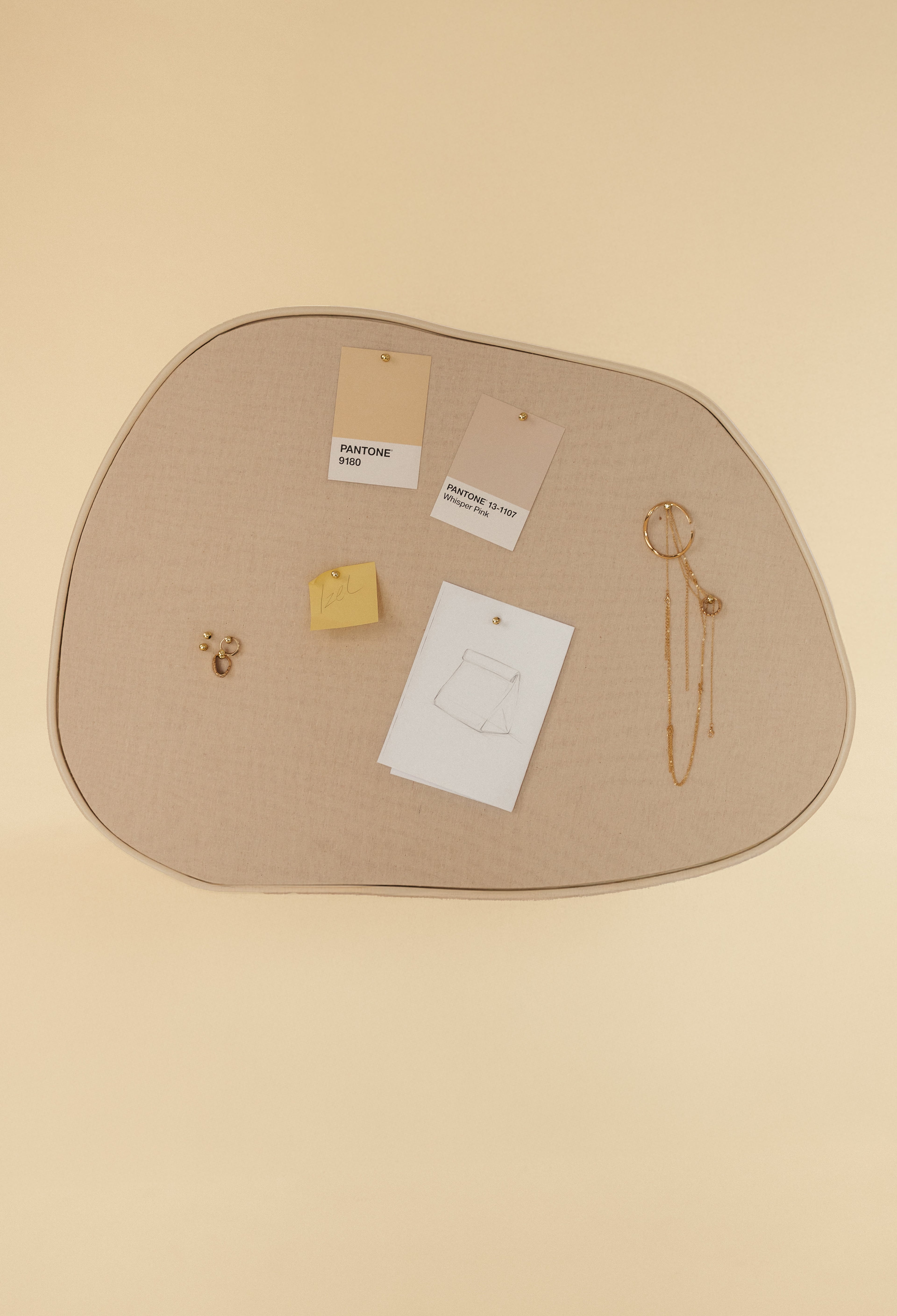 Asymmetrical fabric pinboard with a beige surface and outer frame, displaying pinned Pantone cards, notes, and jewelry against a soft neutral background.