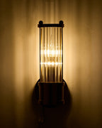 Wall-mounted brass sconce with a cylindrical clear ribbed glass shade illuminated, casting warm vertical light patterns on a textured wall in a dimly lit interior.