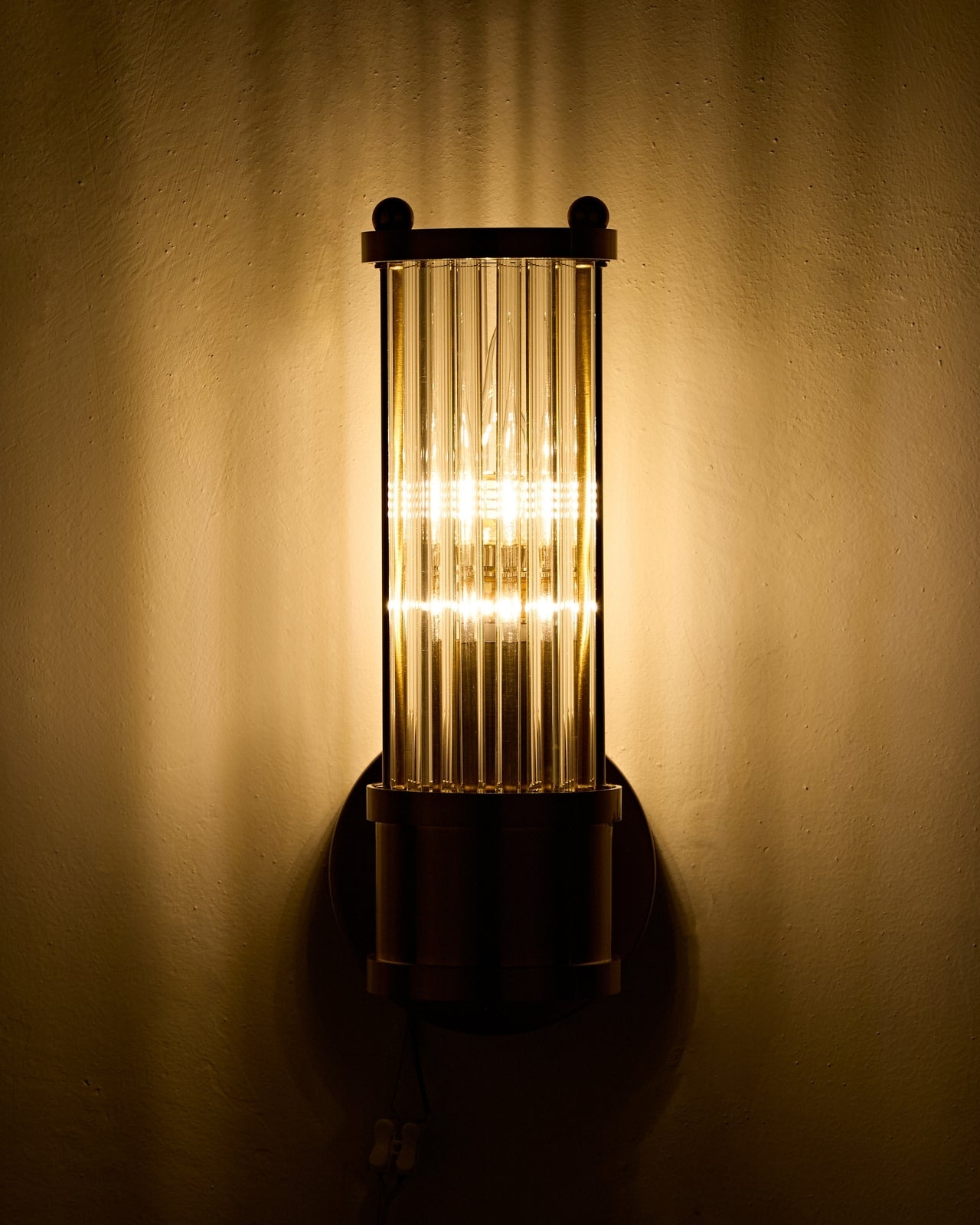 Wall-mounted brass sconce with a cylindrical clear ribbed glass shade illuminated, casting warm vertical light patterns on a textured wall in a dimly lit interior.