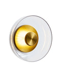 Wall-mounted brass sconce with a circular clear glass shade and a central brass disc, shown lit against a white background, emitting a soft warm light.