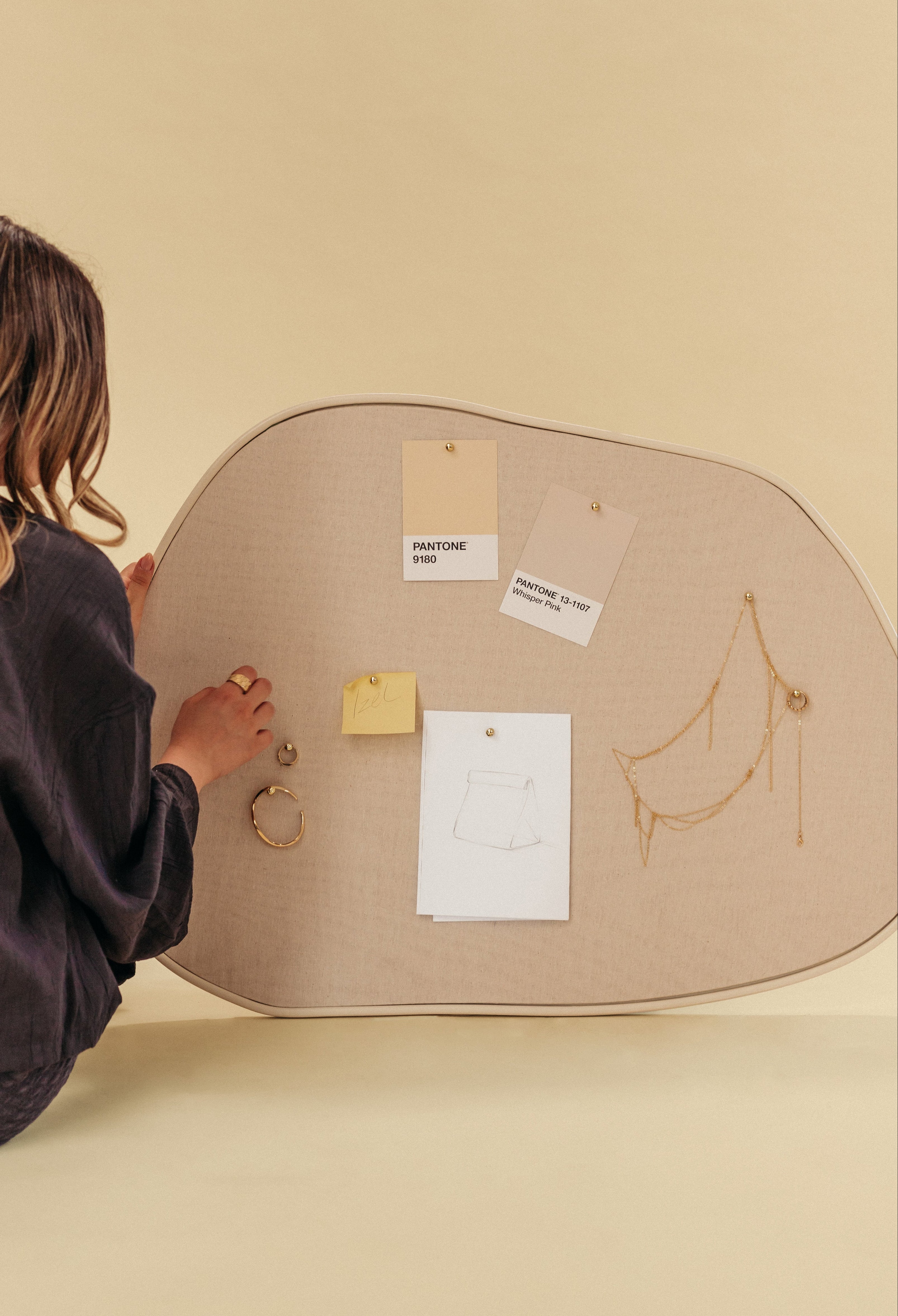A person arranging notes and jewelry on an asymmetrical beige fabric pinboard, set against a soft neutral background.