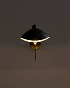 Wall-mounted sconce illuminated on a dark wall, featuring a black dome-shaped metal shade with a white interior, a brass wall plate and vertical stem, and an exposed bulb casting a warm downward glow.