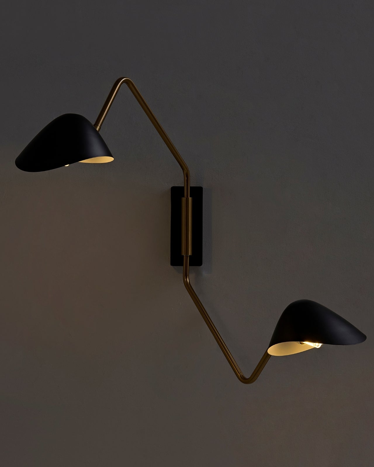 Phineas wall sconce installed on a dark wall, featuring two black metal shades connected by a brass arm, both illuminated and casting warm light upward and downward.