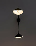 Fleur de Noir Wall Sconce illuminated on a dark wall, featuring a matte black vertical stem with two opposing dome-shaped shades casting a warm glow.