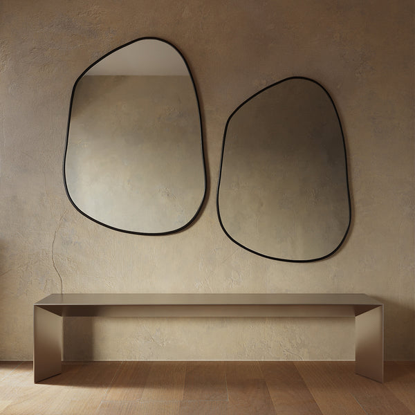Minimalist interior featuring two asymmetrical wall mirrors with thin black frames mounted above a sleek console table, set against a textured beige wall and warm wood flooring.