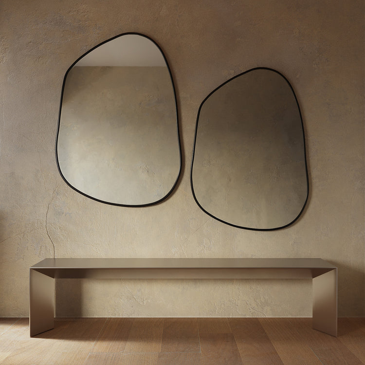 Minimalist interior featuring two asymmetrical wall mirrors with thin black frames mounted above a sleek console table, set against a textured beige wall and warm wood flooring.
