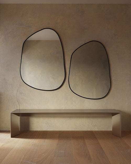 Minimalist interior featuring two asymmetrical wall mirrors with thin black frames mounted above a sleek console table, set against a textured beige wall and warm wood flooring.