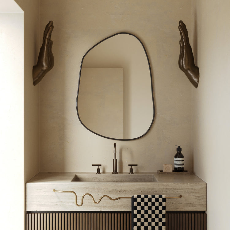 Organic-shaped mirror above a bathroom vanity with a wavy brass towel bar holding a checkered towel.