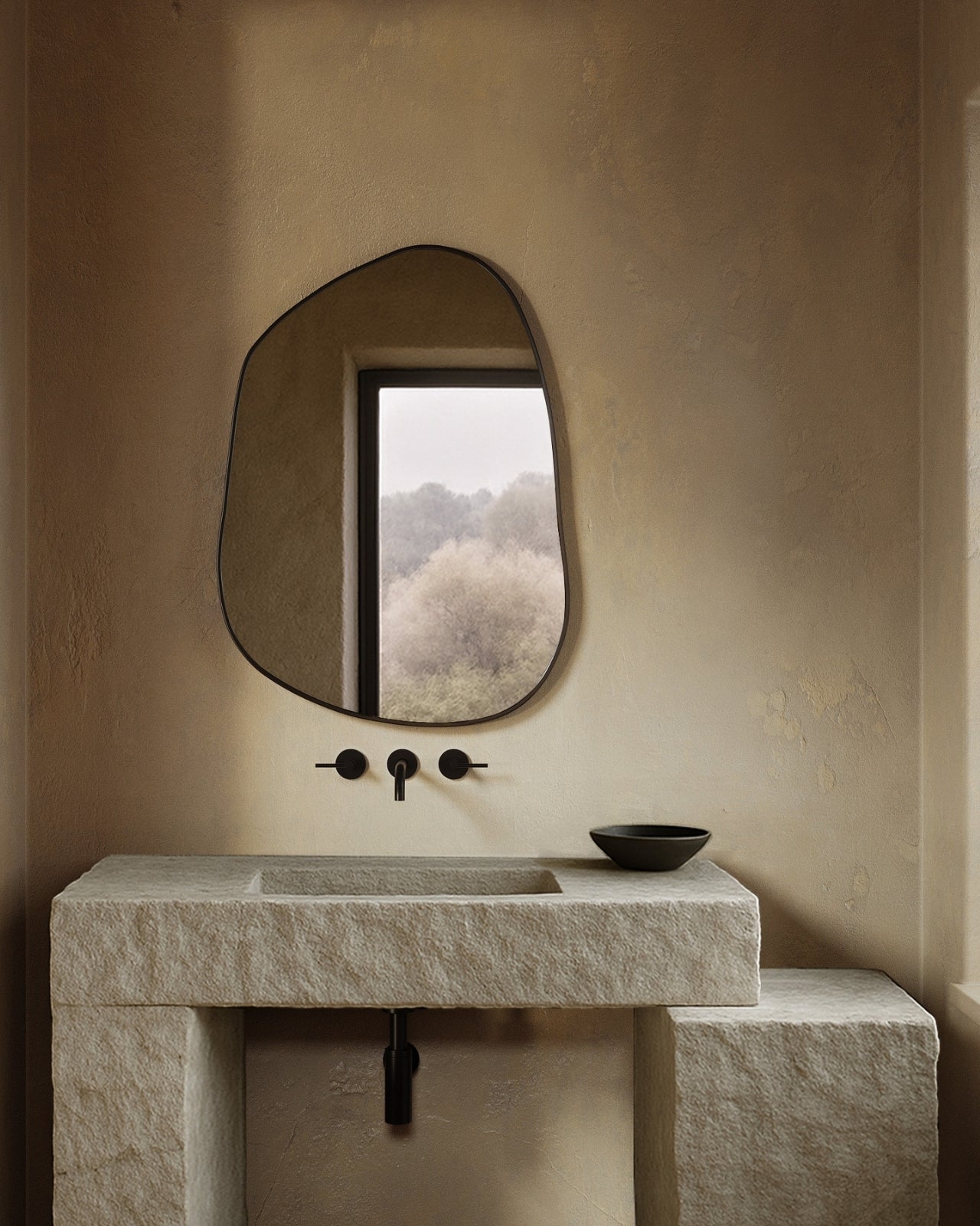Asymmetrical wall mirror mounted above a stone bathroom sink with black fixtures in a minimalist, neutral-toned space.
