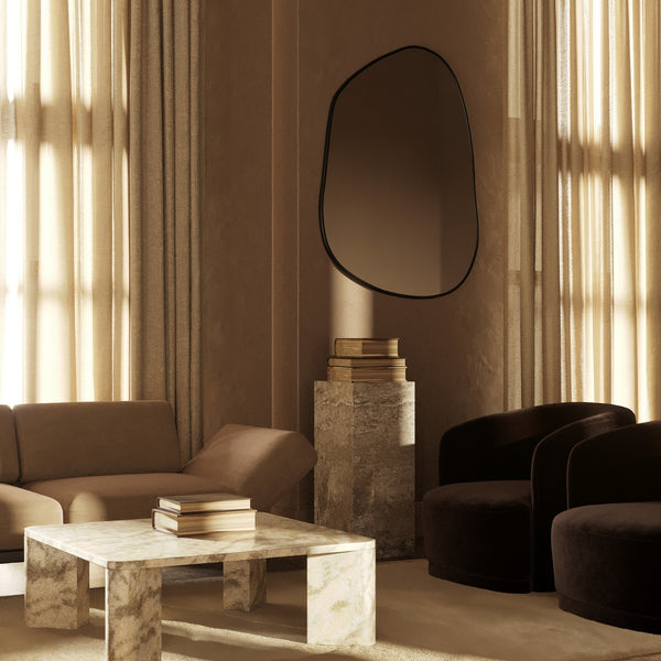 Asymmetrical wall mirror mounted in a warm, neutral-toned living room with stone furniture, soft seating, and filtered natural light.