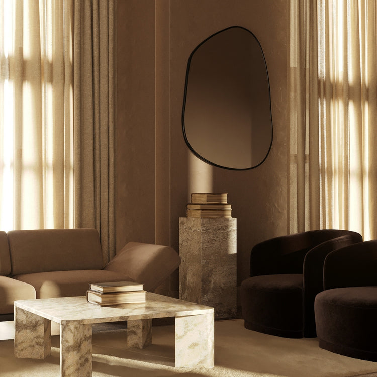 Asymmetrical wall mirror mounted in a warm, neutral-toned living room with stone furniture, soft seating, and filtered natural light.