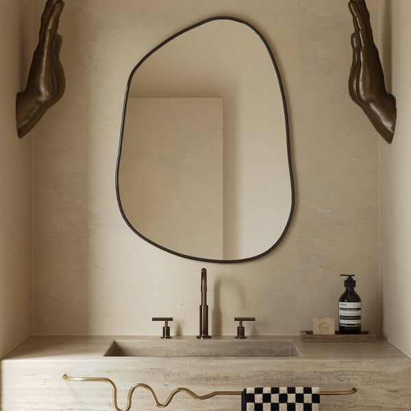 Asymmetrical wall mirror mounted above a stone bathroom vanity with bronze fixtures.