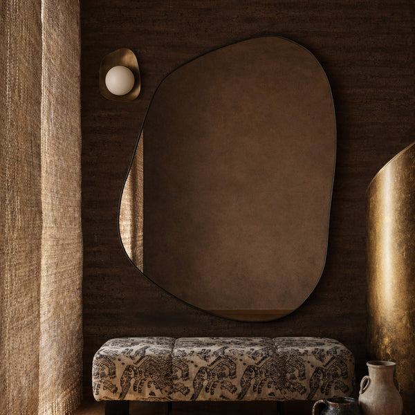 Irregular-shaped wall mirror mounted above a patterned upholstered bench beside decorative vases and sculptural elements.