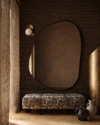 Irregular-shaped wall mirror mounted above a patterned upholstered bench beside decorative vases and sculptural elements.