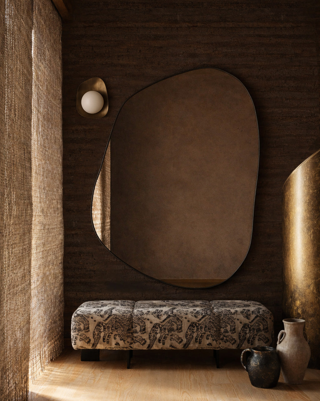 Irregular-shaped wall mirror mounted above a patterned upholstered bench beside decorative vases and sculptural elements.