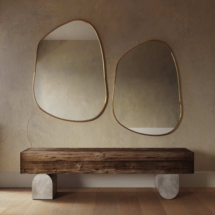 Two asymmetrical mirrors with organic shapes and thin brass-colored frames mounted side by side above a wooden bench.