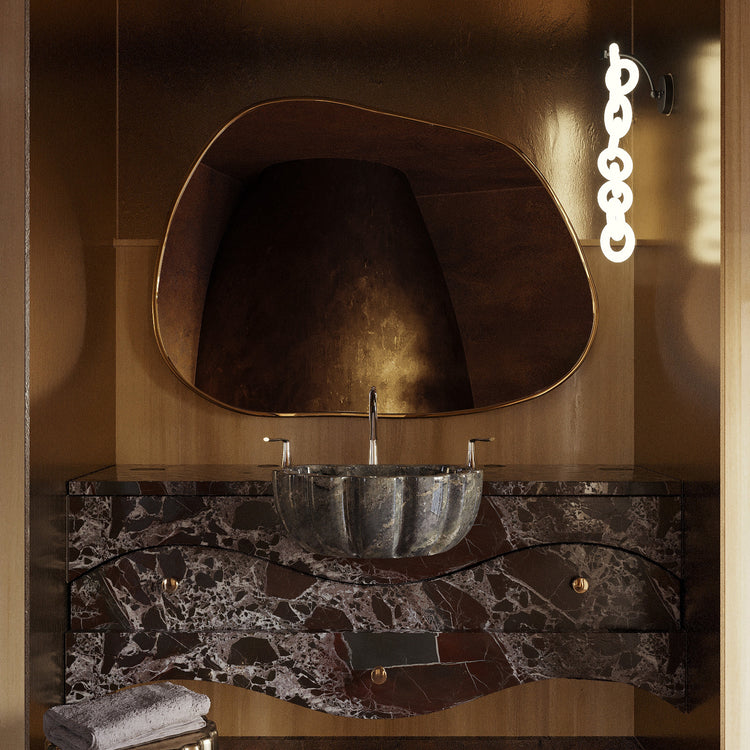 Asymmetrical brass-framed mirror mounted above a marble bathroom vanity with a vessel sink and wall light.