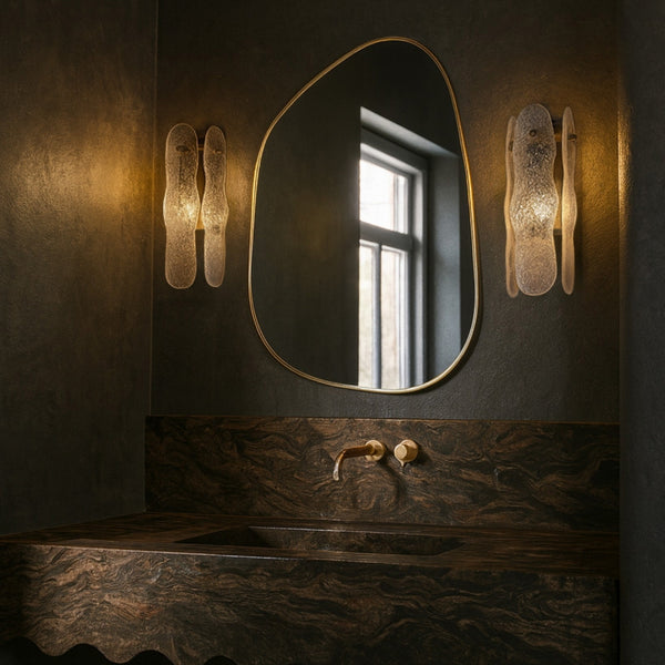 Asymmetrical mirror with an organic shape and a thin brass-colored frame mounted above a bathroom sink, flanked by two wall sconces.