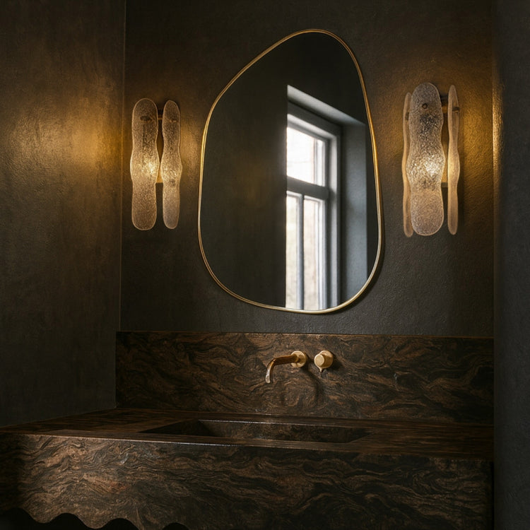 Asymmetrical mirror with an organic shape and a thin brass-colored frame mounted above a bathroom sink, flanked by two wall sconces.