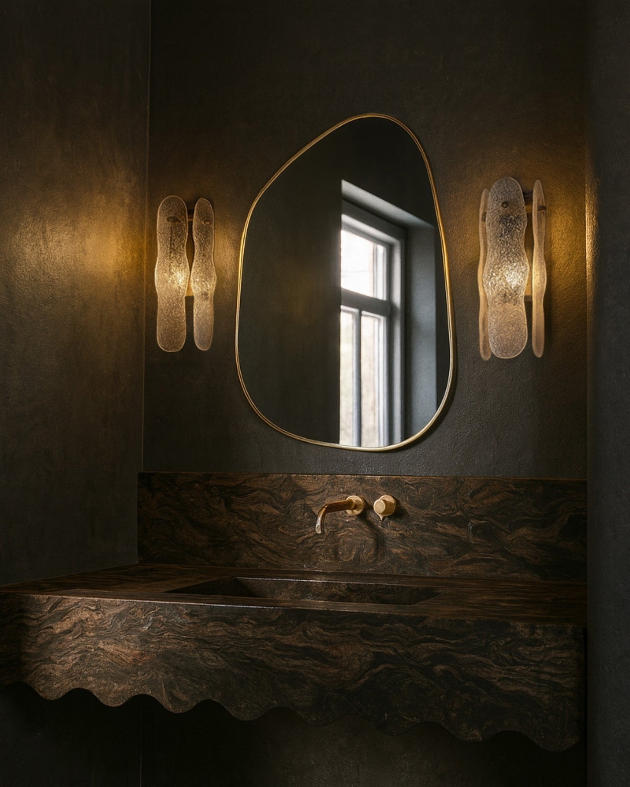 Asymmetrical mirror with an organic shape and a thin brass-colored frame mounted above a bathroom sink, flanked by two wall sconces.