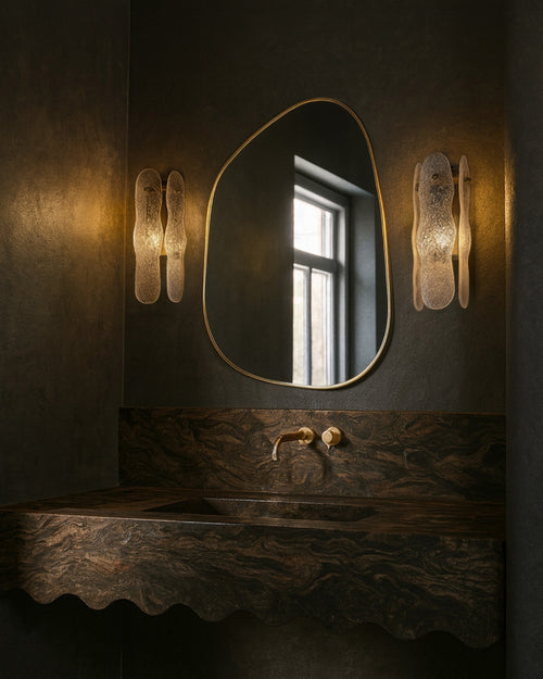 Asymmetrical mirror with an organic shape and a thin brass-colored frame mounted above a bathroom sink, flanked by two wall sconces.