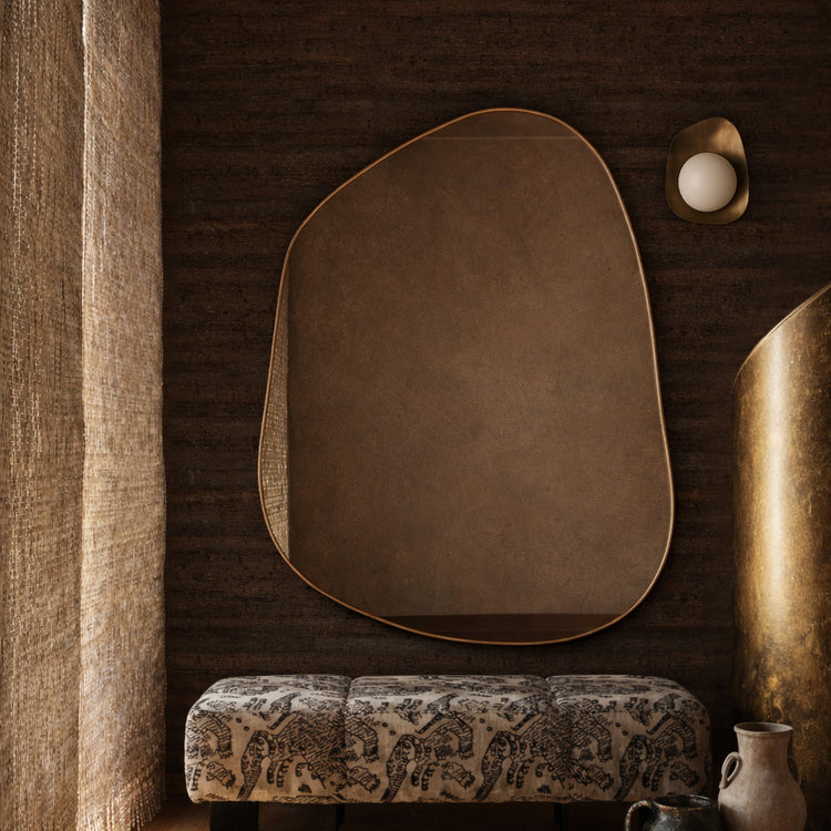 Irregular-shaped wall mirror mounted above a patterned upholstered bench beside decorative vases and sculptural elements.