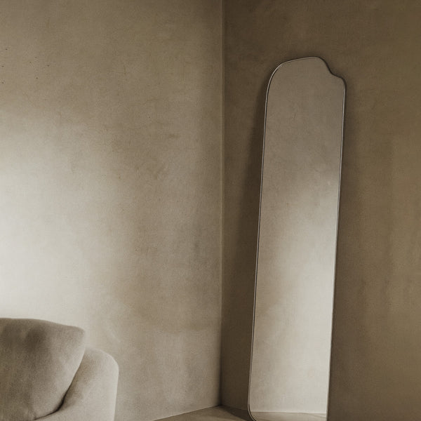 Asymmetrical full-length mirror with an organic shape and thin frame, leaning against the wall in a living room corner with soft natural light and neutral-toned furnishings.