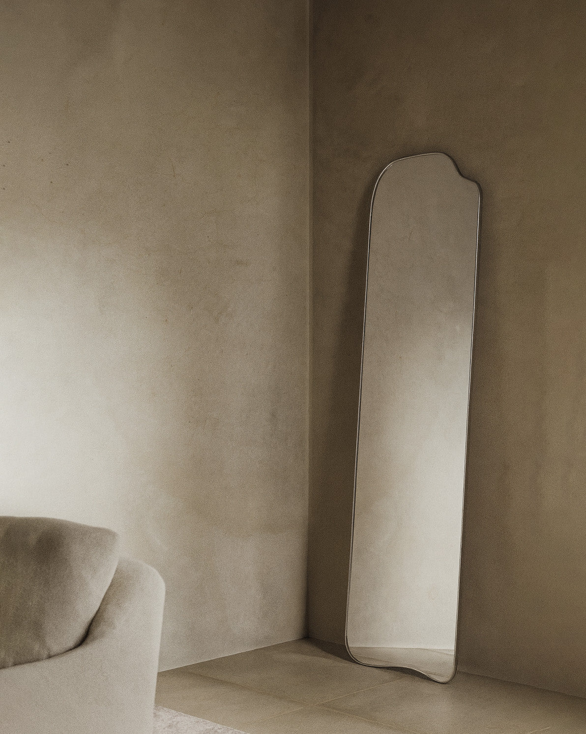 Asymmetrical full-length mirror with an organic shape and thin frame, leaning against the wall in a living room corner with soft natural light and neutral-toned furnishings.