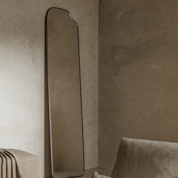 Asymmetrical full-length mirror with an organic shape and thin dark frame, leaning against a textured wall in a living room with soft natural light and neutral upholstered seating nearby.