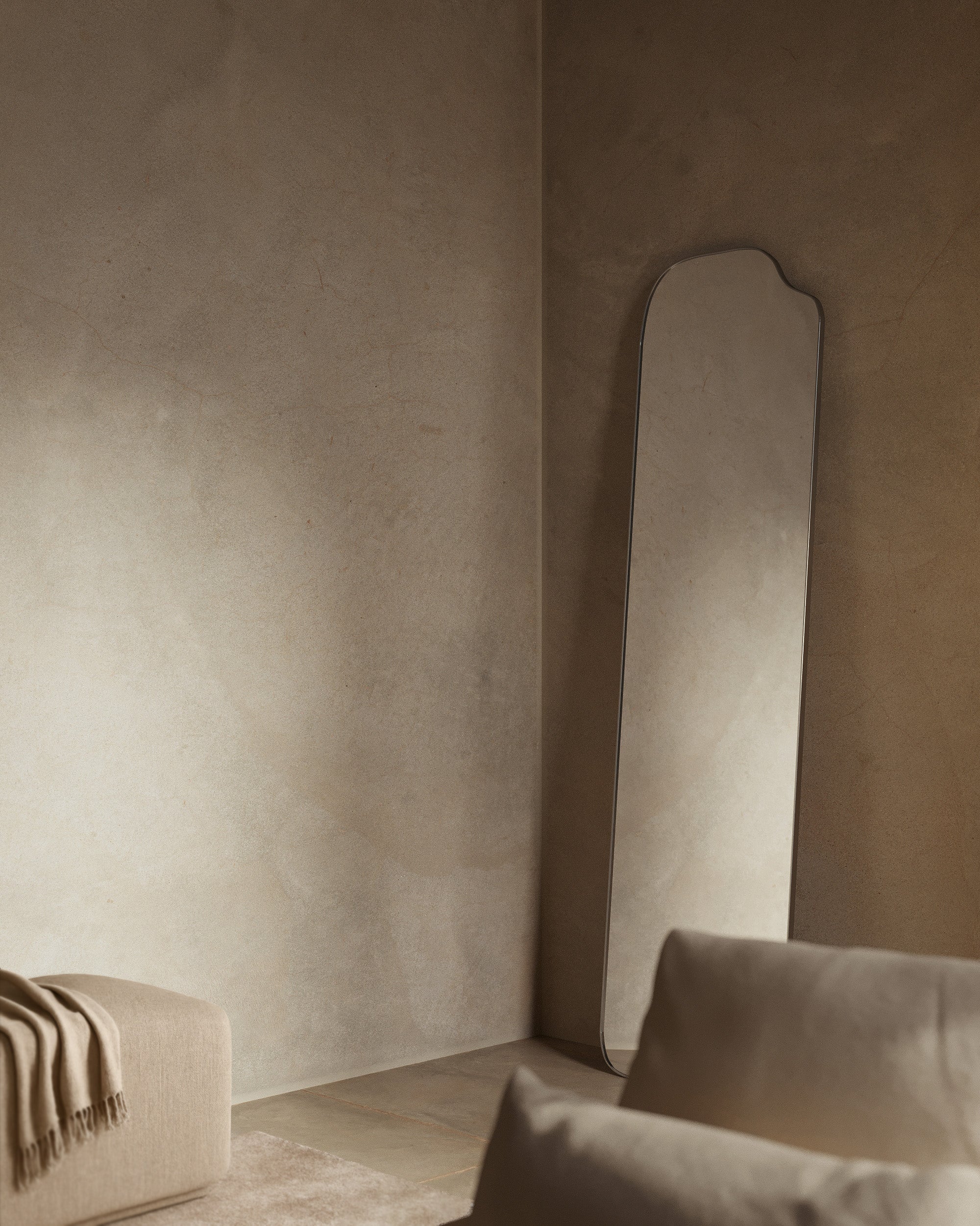 Asymmetrical full-length mirror with an organic shape and thin dark frame, leaning in the corner of a living room as soft natural light falls across textured walls and neutral seating.