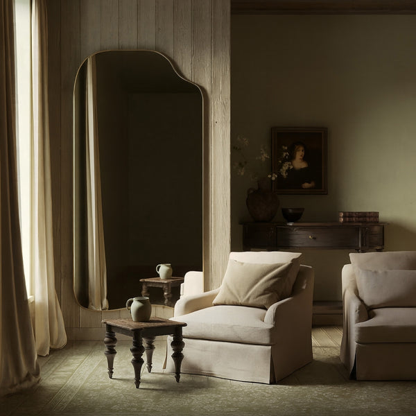 Asymmetrical full-length mirror with an organic shape and thin brass-colored frame, styled in a classic living room with upholstered armchairs, wood paneling, and soft natural light.
