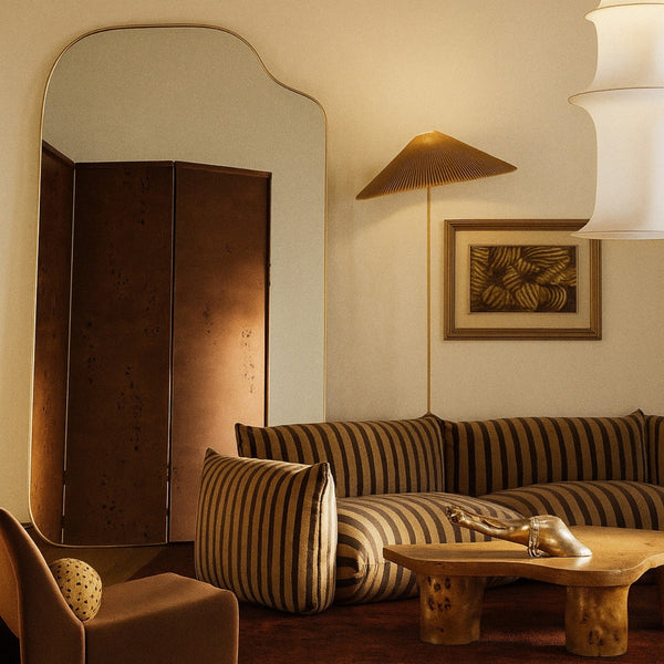 Asymmetrical full-length mirror with an organic shape and thin brass-colored frame, styled in a warm living room with striped seating, sculptural furniture, and ambient lighting.