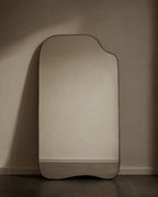Asymmetrical full-length mirror with an organic shape and thin black frame, shown standing against a neutral wall on the floor.