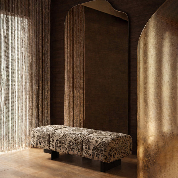 Warm, moody interior with an irregular full length mirror, patterned upholstered bench, textured drapery, and a curved metallic accent panel.