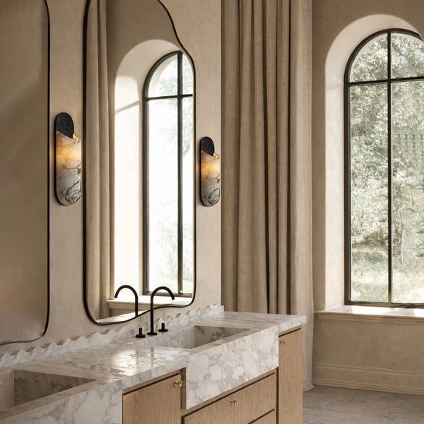 Modern bathroom with a tall, irregular mirror, marble double vanity, black faucets, wall sconces, and large arched windows with beige curtains.
