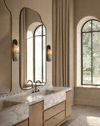 Modern bathroom with a tall, irregular mirror, marble double vanity, black faucets, wall sconces, and large arched windows with beige curtains.